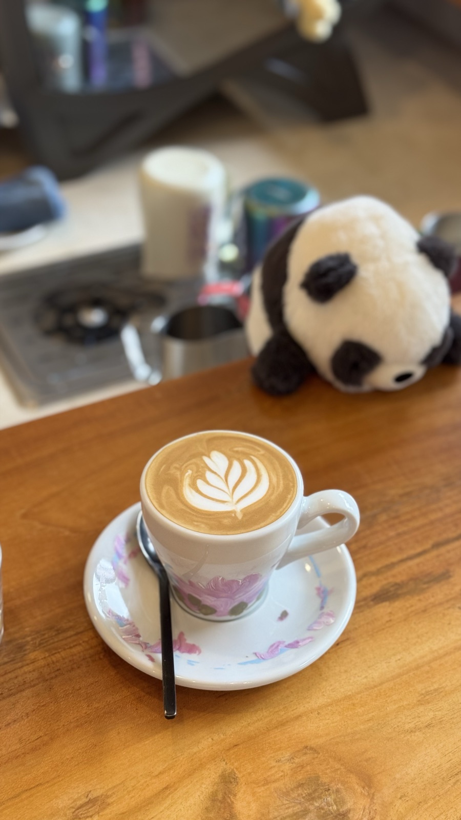 A latte with leaf latte art in a hand-painted cup, with a panda plush behind it